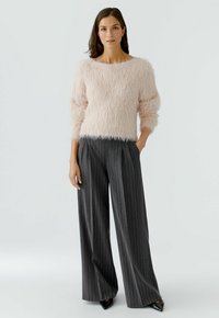 Vrouw in een lichtroze fuzzy trui en donkergrijze gestreepte wijde broek, staand met één hand in haar zak tegen een effen achtergrond.