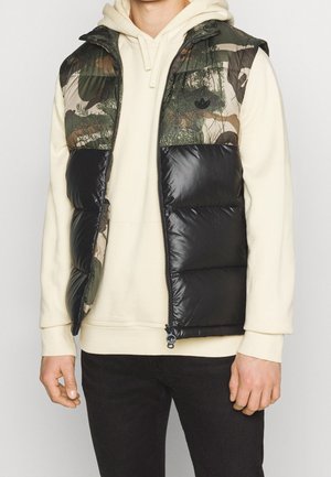 Mężczyzna ubrany w beżową bluzę z kapturem i czarną pikowaną kamizelkę w zielono-brązowy wzór kamuflażu, stojący na jednolitym tle.