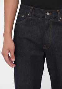 Gros plan sur une personne portant un jean en denim bleu foncé et un haut noir, avec des coutures visibles et une main détendue le long du corps.
