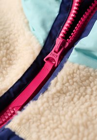 Veste en polaire avec un panneau supérieur bleu clair, un corps en fourrure crème et une fermeture éclaire rose vif. Les couleurs d'accent comprennent le bleu marine et le rouge.