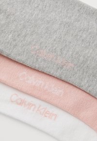 Three pairs of cotton socks in gray, pink, and white, featuring the "Calvin Klein" logo in light pink stitching. Soft texture and simple design.