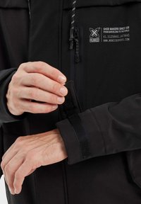 Veste noire avec une fermeture éclair à l'avant et des poignets réglables en Velcro. Présente le logo de la marque et des détails de couture. Le matériau semble lisse et durable.