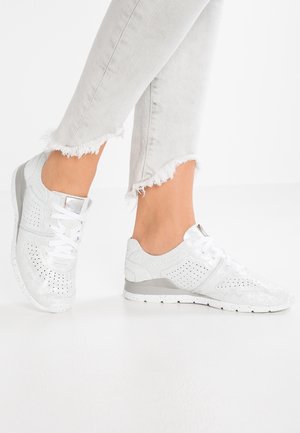 Jambes en jean gris clair à ourlet effiloché portant des baskets basses blanches perforées avec des lacets blancs, debout sur une surface blanche.