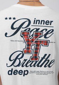Fehér pamut póló, melyen egy grafika található, ami nagy sötétkék betűkkel az "inner Peace" (belső béke) és "Breathe deep" (lélegezz mélyen) feliratokat tartalmazza, valamint piros kockás mintázattal az "YT" betűket.