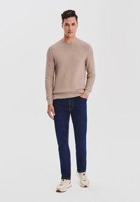 Beżowy sweter dzianinowy o fakturze, okrągły dekolt, zestawiony z ciemnoniebieskimi dżinsami. Model w białych trampkach z dodatkami w innym kolorze. Gładkie tło.