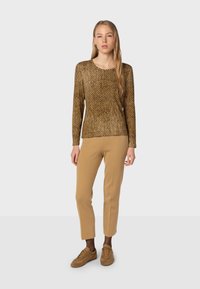 Haut à manches longues, avec un motif marron et or, associé à un pantalon droit beige. Le mannequin porte des baskets marron.