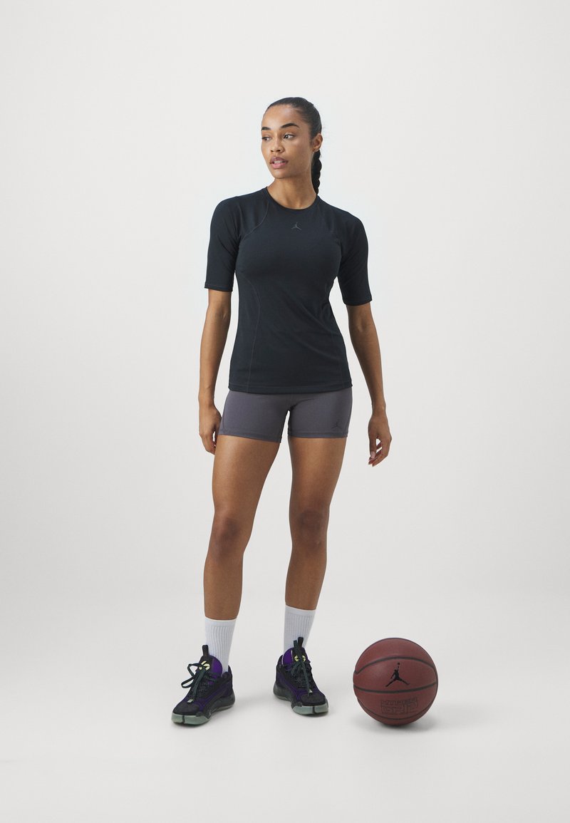 Camiseta deportiva negra de manga corta con detalles grises y un logo, combinada con pantalones cortos grises. Hay una pelota de baloncesto y zapatillas deportivas negras con toques morados.