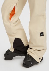 Beige sneeuwbroeken met zwarte accenten, voorzien van zijdelingse ventilatieopeningen met oranje gaas en versterkte boorden. Draagt zwart skischoenen.