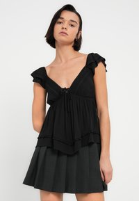 Μαύρο τοπ με κοντά μανίκια και λαιμόκοψη V, με διακοσμητικό δέσιμο μπροστά, στρωματοποιημένες βολάν και υφασμάτινη υφή, συνδυασμένο με πλισέ μαύρη φούστα.