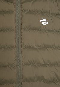 Polstret olivgrønn jakke med vertikale quilted seksjoner, hel glidelås, og en hvit logo på venstre side. Myk, glatt tekstur.