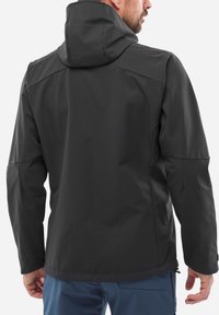 Man wearing a black hooded jacket and blue pants, shown from the back against a plain white background.