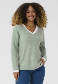 Maglione verde chiaro a trama con scollatura a V, caratterizzato da maniche lunghe e dettagli bianchi sul colletto interno, abbinato a jeans blu.