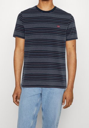 Man draagt een navyblauw en grijs gestreept kort mouw T-shirt met een rood Levi's-logo en lichtblauwe spijkerbroek tegen een effen achtergrond.