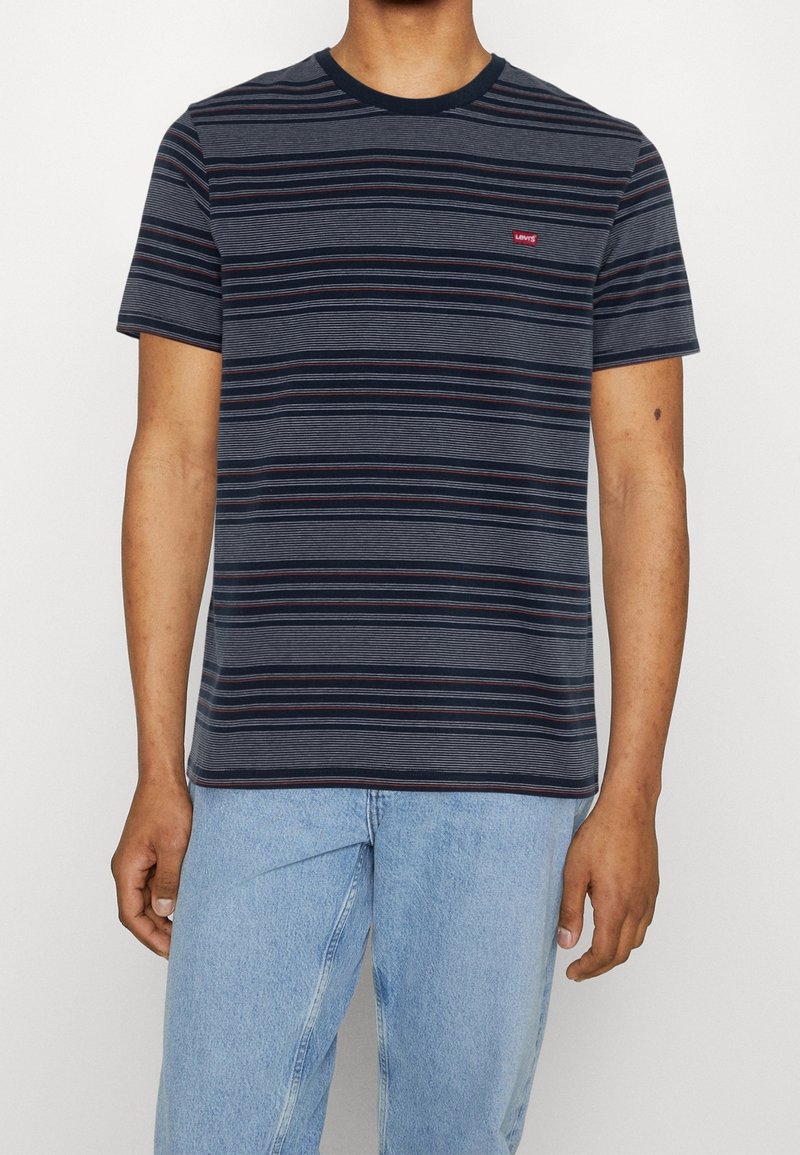 Homme portant un t-shirt à manches courtes rayé bleu marine et gris avec logo Levi's rouge et un jean bleu clair, sur un fond uni.