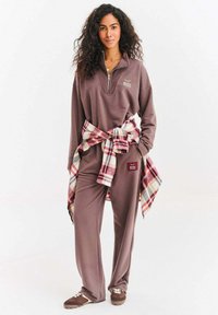 Kobieta o kręconych włosach, ubrana w brązowy dres i trampki, z koszulą w kratę zawiązaną wokół talii, stoi na tle jednolitego białego tła.