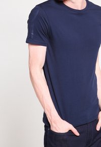 Námořnická modrá bavlněná tričko s krátkým rukávem, kulatým výstřihem a jemným texturovaným detailem na rameni. Hladká tkanina s přiléhavým střihem.