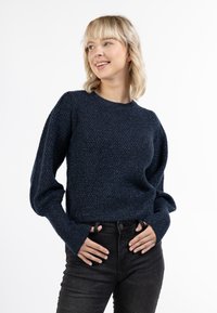 Tengerészkék kötött pulóver texturált mintával, bordás mandzsettákkal és hemmel, valamint klasszikus kerek nyakkivágással, magas derekú sötét farmernadrággal párosítva.