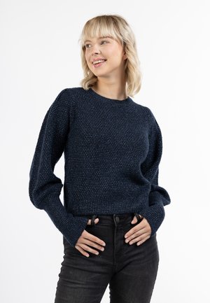 Tengerészkék kötött pulóver texturált mintával, bordás mandzsettákkal és hemmel, valamint klasszikus kerek nyakkivágással, magas derekú sötét farmernadrággal párosítva.