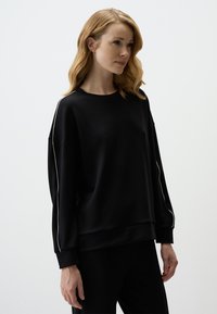 Zwarte sweatshirt met een ontspannen pasvorm, voorzien van een ronde halslijn en witte zijkanten langs de mouwen. Gemaakt van zachte stof met een gladde textuur.