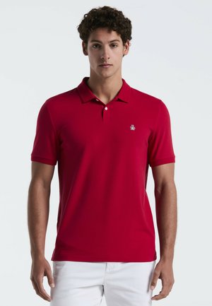 Junger Mann mit lockigem Haar, der ein rotes Poloshirt mit kurzen Ärmeln und weiße Hose trägt, steht vor einem einfarbigen hellen Hintergrund.