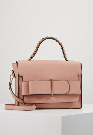Sac à main en cuir rose blush avec un grand nœud sur le devant, une anse chaîne dorée et une bandoulière amovible, posé sur une surface blanche.
