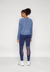 Maglione in misto cotone blu con polsini a coste, abbinato a leggings navy con pannelli in rete e design aderente. Scarpe sportive bianche completano il look.