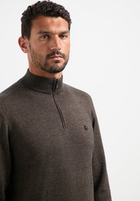 Bruine ritstrui in een gestructureerde brei, met een hoge kraag en een klein logo accent op de linker borst. Subtiel patroon door de stof heen.