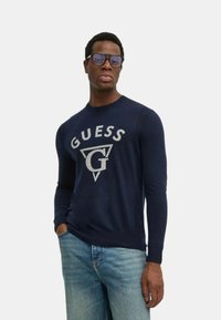 Maglione lavorato a maglia blu navy con un grande logo "GUESS" grigio chiaro, scollo rotondo, dettagli a costine sulle spalle e maniche lunghe.