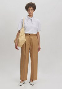 Witte shirt met korte mouwen en een knoopkraag, beige high-waisted broek, beige slouch bag en witte schoenen met zwarte accenten. Effen achtergrond.