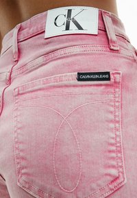Vaqueros de mezclilla rosa con una textura desgastada, que cuentan con una etiqueta blanca que dice "CALVIN KLEIN JEANS" y una etiqueta negra en el bolsillo trasero.