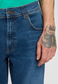 Jeans in denim blu con cuciture arancioni, dotati di chiusura a bottone e tasche anteriori. Tatuaggio visibile sulla mano che tiene il tessuto.