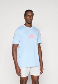 Jasnoniebieska bawełniana koszulka z krótkim rękawem, z różowym logo Adidas i tekstem z przodu. Prosty okrągły dekolt, luźny fason.