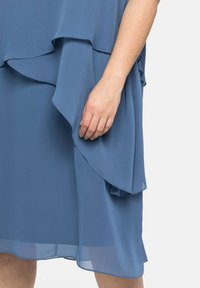 Robe en chiffon bleu à plusieurs couches avec un design fluide ; comporte un ourlet froncé et une texture transparente, partiellement montrée avec une main tenant le tissu.