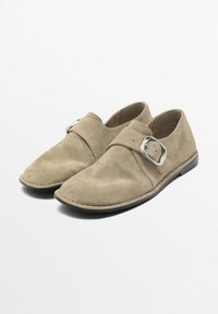Beige suede slip-on shoes with a rounded toe, a side buckle detail, and a flat black sole. Soft texture with seamless construction.