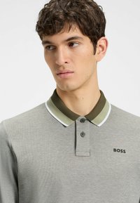 Graues Poloshirt aus strukturiertem Stoff, mit einem dunkelgrünen Kragen, der weiße Akzente aufweist, und zwei Knöpfen. Markenlogo "BOSS" sichtbar.