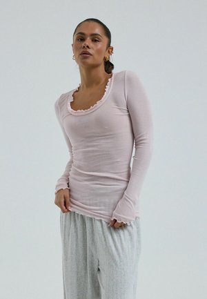 Tumejuustega naine, kellel on hobusesaba, kannab heleroosat pikakarvalist pluusi volangiliste äärtega ja heledaid hallid lahtiseid pükse, seisab ühevärvilise tausta ees.