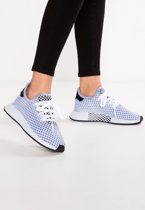 Osoba nosząca niebiesko-białe siateczkowe trampki z białymi sznurówkami i czarnymi akcentami, w zestawie z dopasowanymi czarnymi spodniami na białej podłodze.