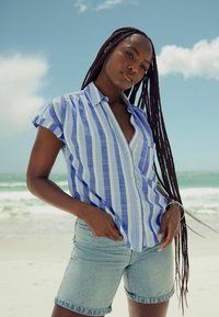 Kurzärmliges Button-up-Hemd mit blauen und weißen vertikalen Streifen, kombiniert mit hellblauen Jeansshorts, vor einem Strandhintergrund.