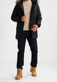 Svart parkajacka med foder av päls i huvan, dragkedja och tryckknappsstängning, bärs över en beige tröja, kombineras med mörka jeans och bruna stövlar.