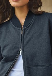 Donkergrijze bomberjack met geribbeld kraag en manchetten, voorzien van een zilveren ritssluiting en een gladde stofstructuur. Witte shirt zichtbaar eronder.