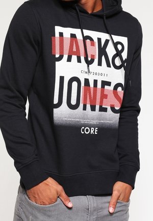 Sort hættetrøje med hvid og rød bloktekst, der siger "JACK & JONES CORE", båret af en person med hænderne i lommerne på grå bukser.