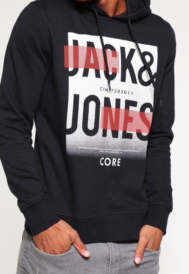 Sweat à capuche noir avec texte en blocs blancs et rouges indiquant "JACK & JONES CORE" porté par une personne les mains dans les poches d'un pantalon gris.