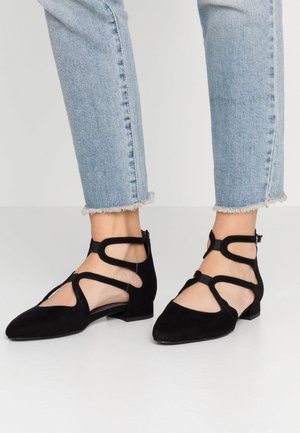 Fødder iført sorte sandal-flade sko med spids snude, parret med lyseblå jeans med frynset kant mod en ensfarvet baggrund.