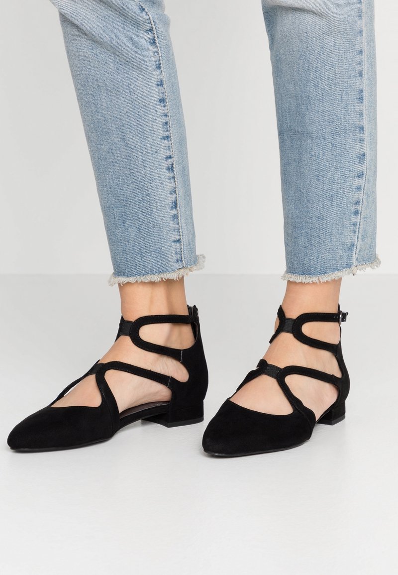 Pieds portant des ballerines noires à brides avec bout pointu, associées à un jean bleu clair à ourlet effiloché sur un fond uni.