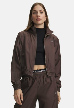 Vrouw draagt een bruine jas met rits en bijpassende broek met een zwart cropped topje eronder, staand tegen een effen achtergrond.