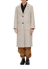 Hellgrauer Wollmantel mit Kragen, zwei großen vorderen Taschen und schwarzen Knöpfen. Getragen über einem schwarzen Hemd und braunen Hosen, kombiniert mit schwarzen Stiefeln.