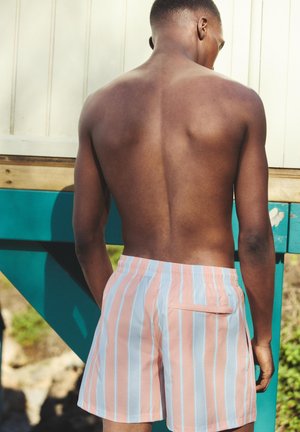 Homme torse nu debout à l'extérieur, de dos, portant un short à rayures verticales roses et bleu clair, près d'une structure en bois bleu sarcelle et blanc.