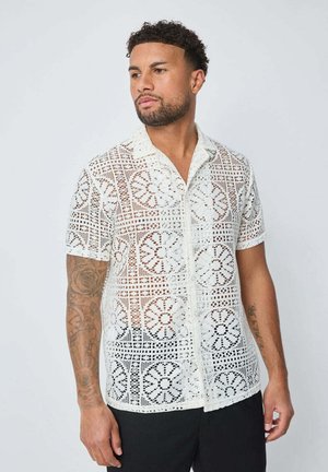 Homme aux cheveux courts et bouclés avec des tatouages sur les deux bras porte une chemise blanche courte en crochet et un pantalon noir, regardant sur le côté.