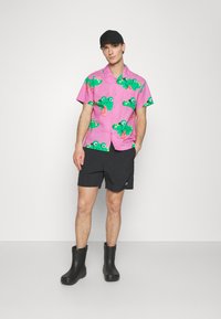 Camisa de mangas curtas rosa com padrões de sapos verdes, combinada com calções pretos e botas de borracha pretas. Design simples, textura casual.