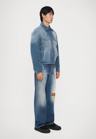 Male model wearing faded blue denim jacket and wide-leg jeans with patch details, standing against plain white background.